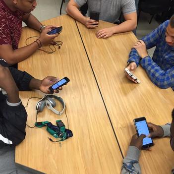 A photo of classroom table, taken from above. Sitting at the table are five students, all actively on their phones. One student has over ear headphones on the table beside him, another has ear buds tangled in front of him. One phone is angled as if taking a photo of the person taking this photo. 